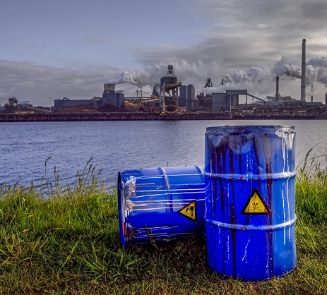 Empty Blue Chemical Waste Drums Lying on an Abandoned Bank with a view on Smoking Exhaust Pipes of a Heavy Industrial Factory